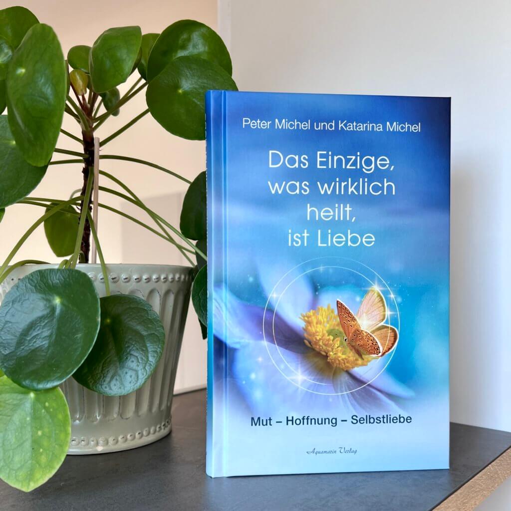 Das Bild zeigt das Buch "Das Einzige, was wirklich heilt, ist Liebe" mit einem blauen Cover vor einer grünen Zimmerpflanze.