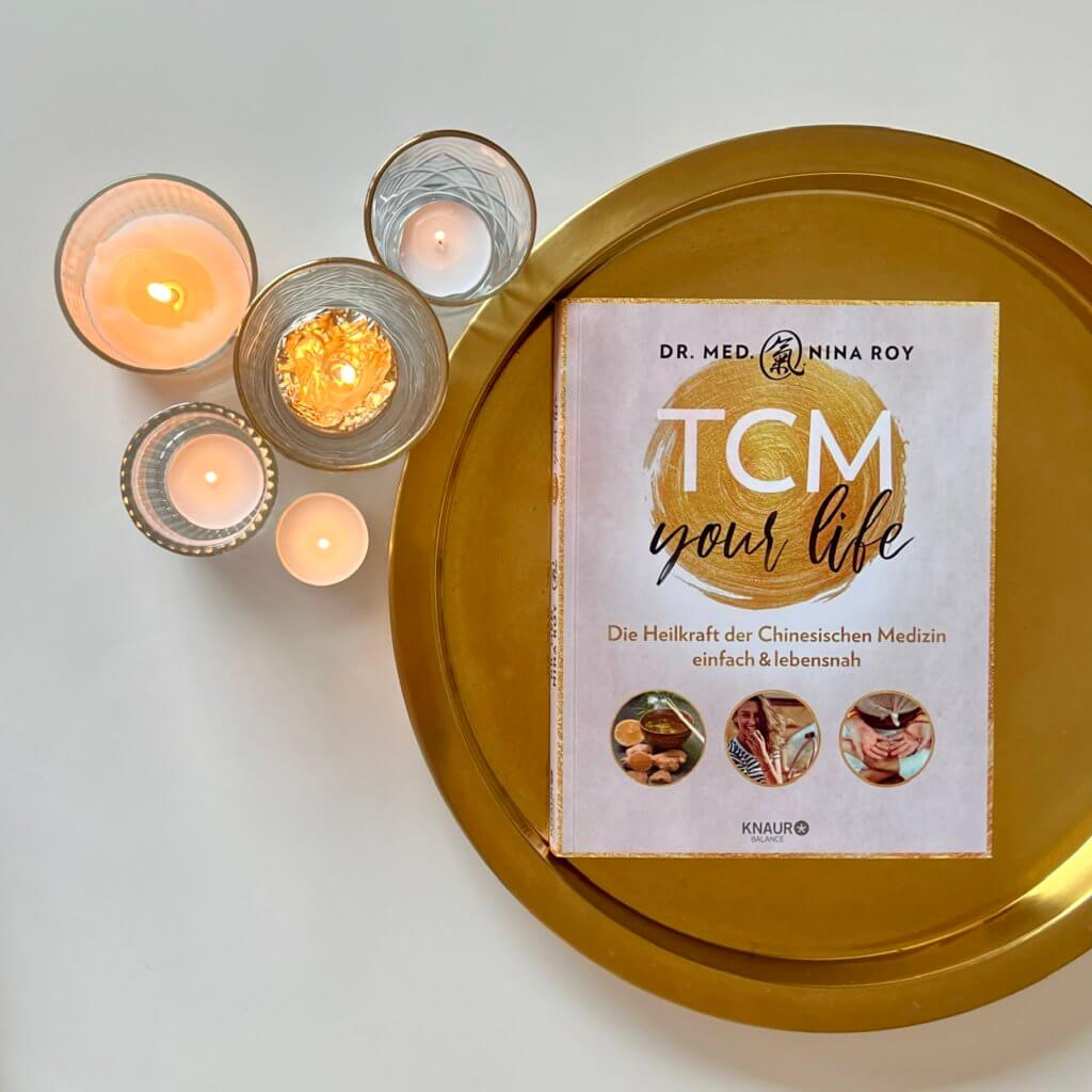Das Bild zeigt das Buch "TCM your life" von Dr. med. Nina Roy auf einem goldenen Tablet neben Kerzen liegend.