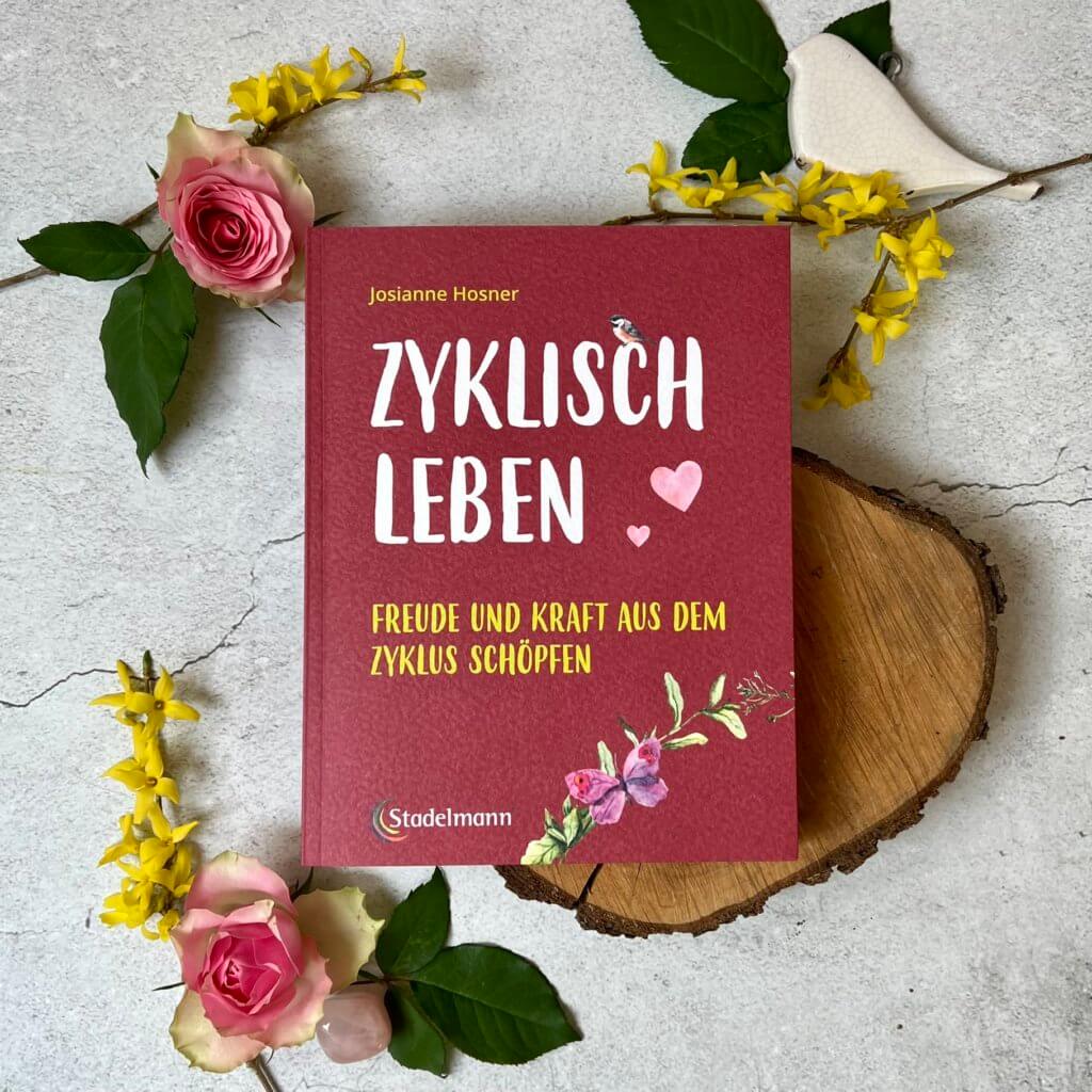 Das Bild zeigt das Buch "Zyklisch leben" von Josianne Hosner umgeben von Blumen auf grauem Hintergrund.