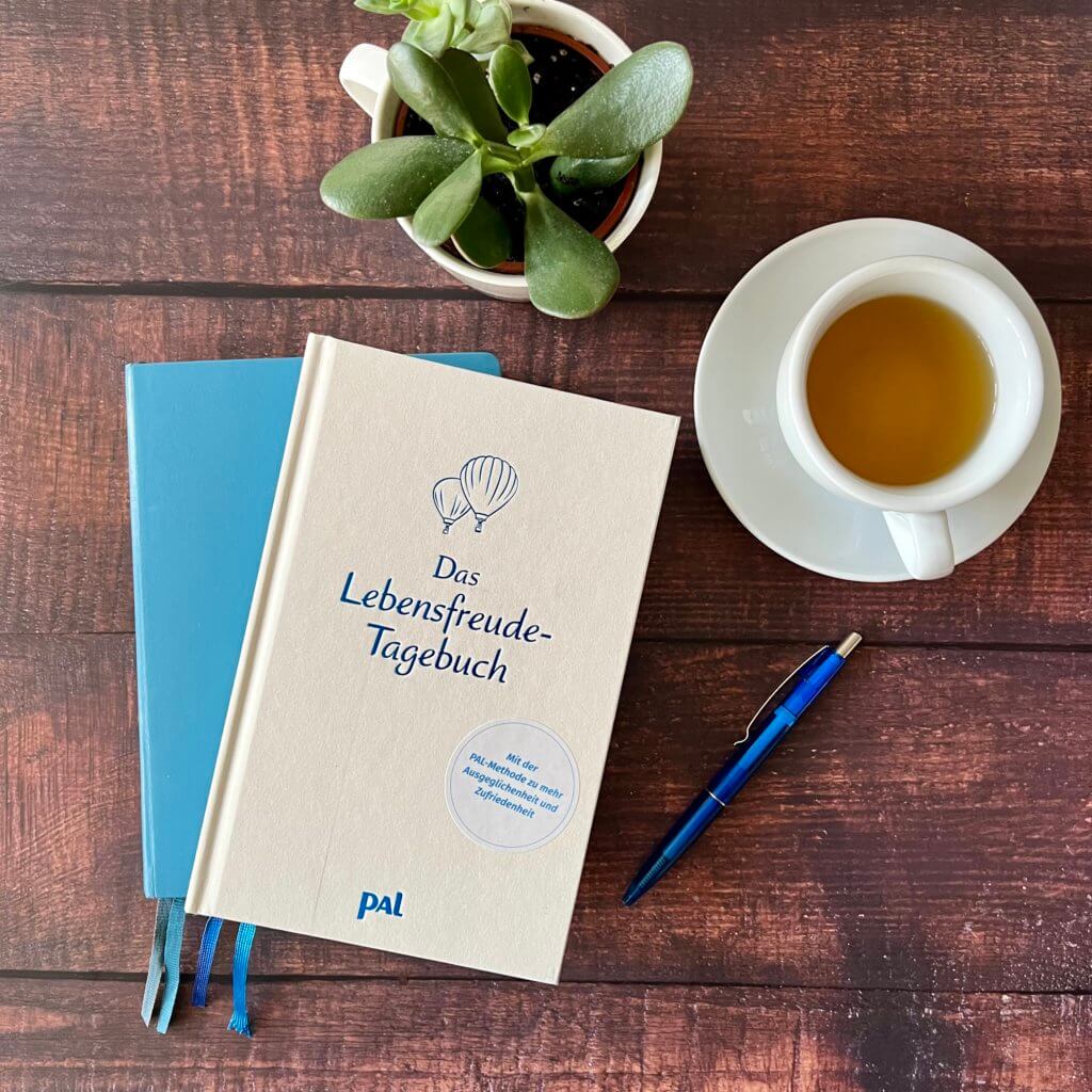 Das Bild zeigt das "Lebensfreude-Tagebuch" auf einem blauen Notizbuch liegend, umgeben von Tee und einer Pflanze.
