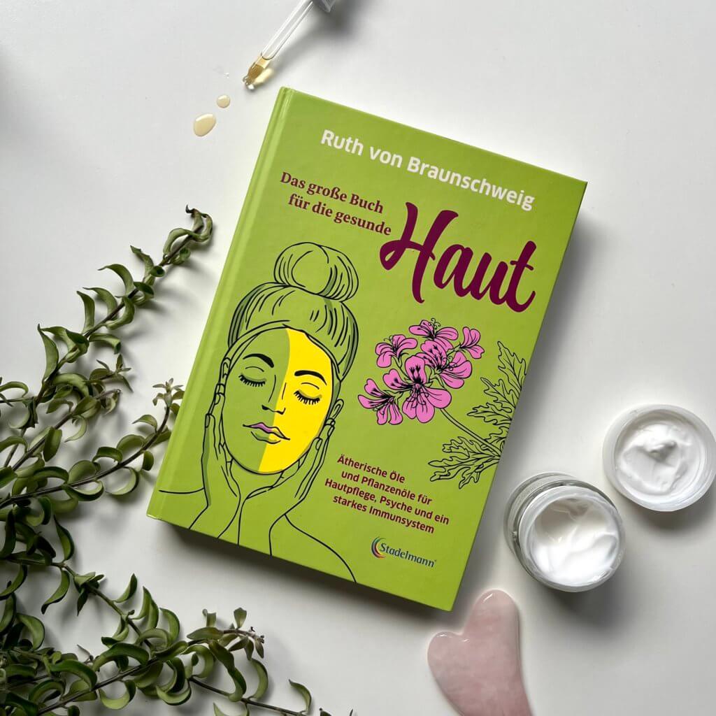 Das Bild zeigt das Buch "Das große Buch für die gesunde Haut" von Ruth Braunschweig umgeben von einer Pflanze, einem Gua Sha und einer Gesichtscreme.